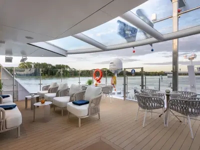 Eine moderne, überdachte Panorama-Lounge auf der Amadeus Amara mit bequemen Polstersesseln. Durch die Glasfront ist ein Fluss mit bewachsenen Ufern sichtbar. Keine Personen.