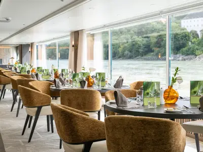Ein elegantes Restaurant auf der AMADEUS Amara mit einer Reihe gedeckter Tische und Stühle. Große Fenster bieten einen weiten Blick auf einen Fluss mit bewaldeten Ufern und einem weißen Gebäude in der Ferne. Die Tische sind mit grünen Menükarten, Gläsern und orangefarbenen Vasen dekoriert.
