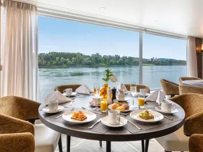 Grosses, helles Restaurant auf der AMADEUS Amara mit Tischen fuer ein Fruehstueck und weitem Blick auf einen Fluss und gruene Ufer. 