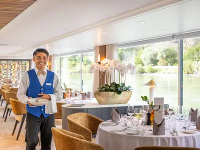 Ein lächelnder Kellner in blauer Weste hält einen Teller in einem eleganten Restaurant auf der Amadeus Amara mit großen Fenstern und Flussblick.