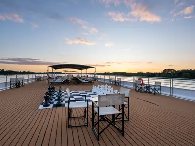 Blick auf das leere Sonnendeck der Amadeus Amara bei Sonnenuntergang. Ein riesiges Schachbrett mit schwarzen und weissen Figuren steht im Vordergrund. Dahinter sind Liegestühle und eine überdachte Sitzgruppe zu sehen. Der Fluss und bewaldete Ufer bilden den Horizont.