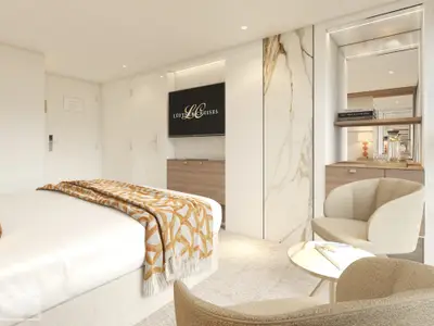 Blick in eine moderne und luxuriöse Kabine der Amadeus Aurea mit einem hellen Doppelbett, gemusterter Decke, Flachbildfernseher mit 'Lüftner Cruises'-Logo, Holzschränken und einer gemütlichen Sitzecke mit Sessel und kleinem Tisch. Eine marmorierte Wandpaneele und ein Spiegelbereich mit Büchern und einer Wasserkaraffe sind ebenfalls zu sehen. Die Beleuchtung ist warm und einladend.