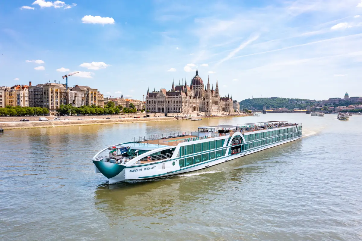 Flusskreuzfahrtschiff AMADEUS BRILLIANT auf dem Fluss, das ungarische Parlament und die Skyline von Budapest bei Tag.