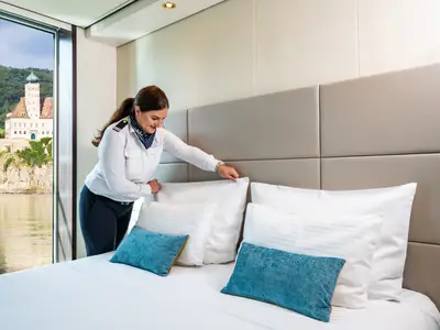 Ein Crew-Mitglied in Uniform macht das Bett in einer modern eingerichteten Flusskreuzfahrtkabine auf der Amadeus Brilliant. Durch das große Fenster ist eine historische Burg auf einem Felsen am Flussufer zu sehen. Die Kabine ist hell und hat blaue Akzente.