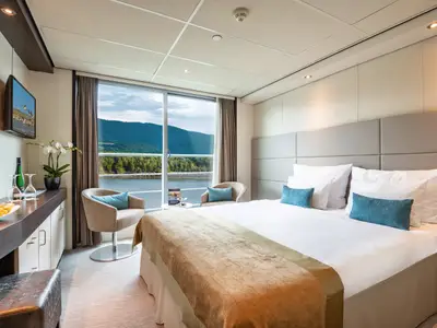 Eine helle und moderne Balkonkabine auf dem Mozart and Strauss Deck der Amadeus Brilliant. Ein bequemes Doppelbett, zwei Sessel und eine grosse Fensterfront mit Blick auf einen Fluss und bewaldete Berge sind zu sehen.