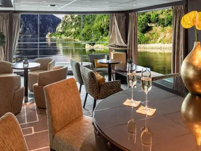 Elegante Lounge auf der Amadeus Brilliant mit bequemen Sesseln und Tischen. Sektgläser und eine Vase schmücken den Vordergrund. Panoramablick auf Fluss und Berge.