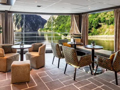 Blick in eine helle, moderne Panorama-Lounge auf der AMADEUS Brilliant mit raumhohen Fenstern. Die komfortablen Sessel und Tische bieten eine entspannte Atmosphäre mit Aussicht auf eine malerische Flusslandschaft mit grünen Berghängen unter blauem Himmel. 