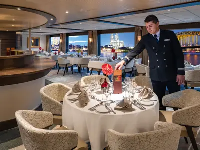 Ein Kellner platziert eine Speisekarte auf einem elegant gedeckten Tisch in einem Restaurant auf der Amadeus Brilliant. Im Hintergrund sind durch grosse Fenster die beleuchtete Kölner Skyline und der Rhein bei Abenddämmerung zu sehen.