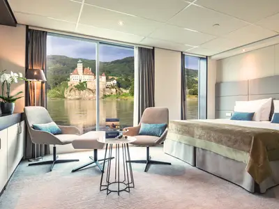 Luxuriöse Flusskreuzfahrtkabine mit großem Bett, Sitzbereich und Panoramafenstern. Blick auf eine historische Burg am Fluss und grüne Hügel. Moderne, elegante Einrichtung mit TV und Orchidee.