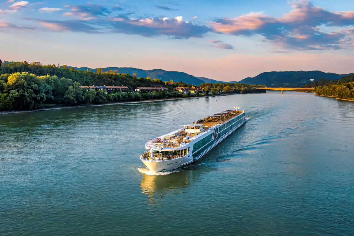 AMADEUS Cara mit vielen Decks und grossen Fenstern, befährt einen Fluss umgeben von gruenen Huegeln und einer Bruecke im Hintergrund waehrend des Sonnenuntergangs.