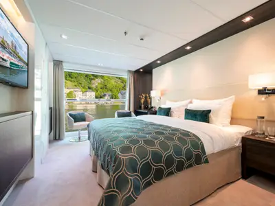 Innenansicht einer hellen und modernen Kabine auf der AMADEUS Cara mit Doppelbett, Fenstern mit Blick auf einen Gruenen Fluss und eine Stadt am Ufer.
