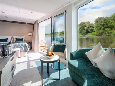 Geräumige und helle Suite auf der AMADEUS Cara mit Doppelbett, Sitzbereich und bodentiefen Fenstern, die einen Blick auf einen grünen Fluss bieten. Eine Flasche Champagner und Obst stehen bereit.