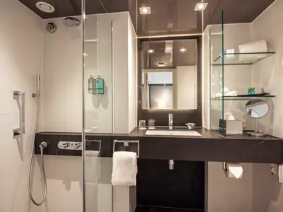 Modernes Badezimmer einer Kreuzfahrtkabine mit weissen Fliesen, dunkler Ablage, grosser Dusche, Waschtisch und Beleuchtung.