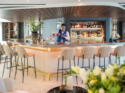 Ein Barkeeper in blauer Weste mixt einen Cocktail an einer eleganten Bar mit Messingakzenten und Hockern. 