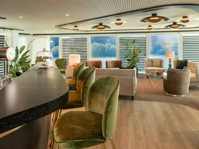 Amadeus Club: Elegante Bar-Lounge auf der AMADEUS Cara. Dunkle Marmortheke, olivgrüne Samtbarhocker und bequeme Loungesessel. Grosse Fensterfront bietet Blick auf eine grüne Flusslandschaft.