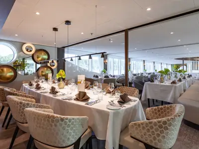 Ein elegantes Restaurant auf der AMADEUS Cara mit einem langen, weiss gedeckten Tisch und bequemen Stuehlen. Der Raum ist hell und hat grosse Fenster mit Blick nach draussen. Mehrere runde Dekoelemente an der Wand schmuecken den Raum. Im Hintergrund sind weitere Tische zu sehen.