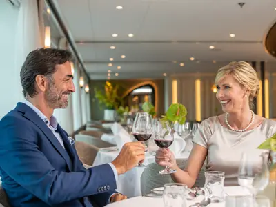 Ein elegant gekleidetes Paar, Mann und Frau, laechelt und stoesst in einem modernen Restaurant auf der AMADEUS Cara mit zwei Gläsern Rotwein an. 