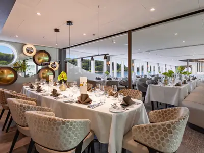 Elegantes Schiffsrestaurant auf der AMADEUS Cara mit gedeckten Tischen. Panoramablick auf Fluss und Wald. Runde Wanddekorationen.