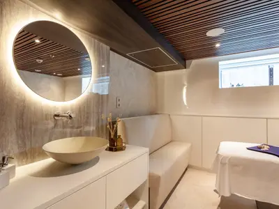 Ein heller Spa-Bereich auf der AMADEUS Cara mit einem ovalen, beleuchteten Spiegel über einem Waschbecken und einer Massageliege. 
