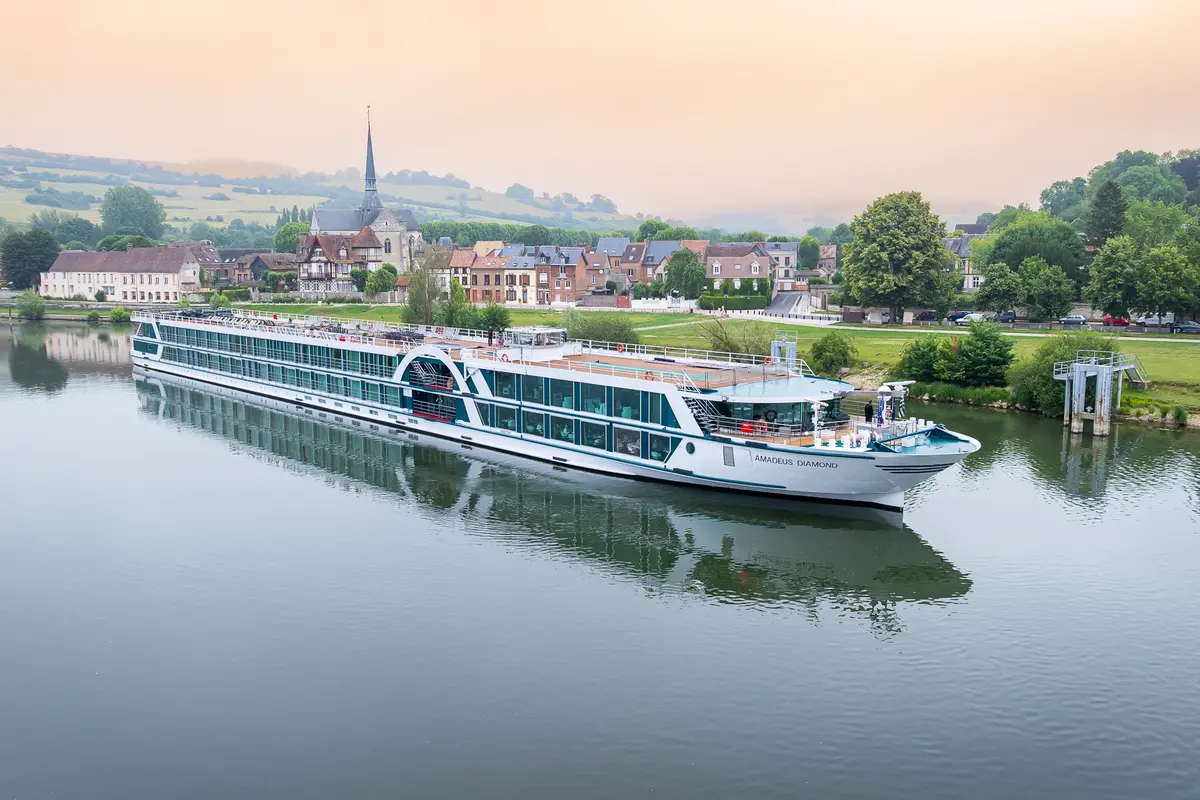 Ein weißes Flusskreuzfahrtschiff namens AMADEUS DIAMOND fährt auf einem ruhigen Fluss. Im Hintergrund sind grüne Hügel und ein malerisches Dorf mit einer Kirche zu sehen. Der Himmel ist hell und leicht bewölkt.