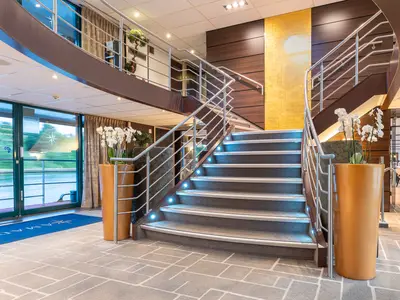 Innenansicht eines modernen Kreuzfahrtschiff-Atriums auf der AMADEUS Diamond mit einer eleganten, geschwungenen Treppe. Die Treppe hat LED-Beleuchtung in den Stufen und Edelstahlgeländer. Große Fenster bieten einen Blick auf eine grüne Uferlandschaft und Wasser. Im Vordergrund stehen große orangefarbene Pflanzgefäße mit weißen Orchideen.