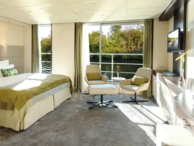 Moderne Suite auf der AMADEUS Diamond. Doppelbett, Sitzbereich mit zwei Sesseln am Panoramafenster. Blick auf Fluss und Bäume. Sideboard mit TV und Champagnerflasche.