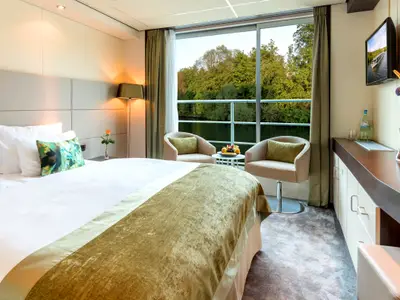 Blick in eine moderne, helle Aussenkabine auf der AMADEUS Diamond. Ein gemachtes Doppelbett mit grünen Kissen ist zu sehen. Am Fenster stehen zwei Sessel mit Blick auf bewaldetes Ufer.