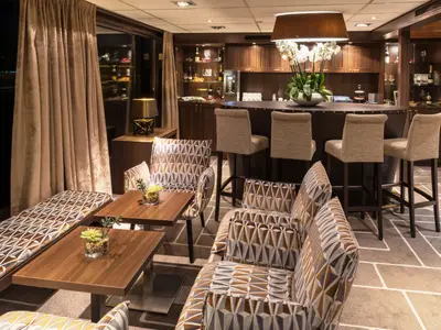 Ein eleganter Barbereich auf der AMADEUS Diamond mit einer dunklen Holztheke, bequemen Barhockern und mehreren Sitzgruppen mit gemusterten Polstern und Holztischen.