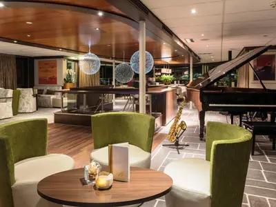 Elegante Innenansicht einer Schiffsbar-Lounge am Abend mit einem schwarzen Flügel und einem goldenen Saxophon. Grüne Sessel und Holztische dominieren den Vordergrund. Die Decke ist aus Holz, während der Boden eine Mischung aus Holz und gemustertem Teppich aufweist. Drei moderne, kugelförmige Lichtinstallationen hängen von der Decke. Im Hintergrund sind weitere Sitzbereiche und große Fenster mit Blick auf die Nacht zu sehen.