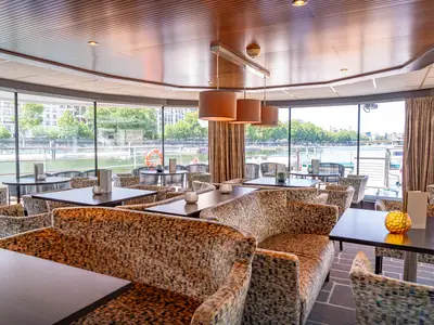 Eleganter Lounge-Bereich auf der AMADEUS Diamond mit gemusterten Sitzmöbeln und Tischen. Grosse Fensterfront bietet Blick auf Fluss und Stadt mit Bäumen.