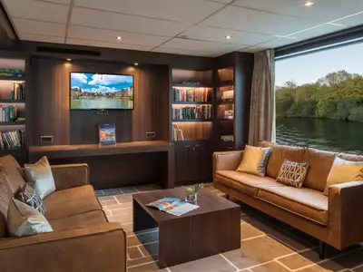 Einladende Bibliothek-Lounge an Bord der AMADEUS Diamond mit bequemen Ledersofas, Holzbüchernregalen, einem Flachbildfernseher und einem großen Fenster mit Blick auf einen grünen Flussufer.