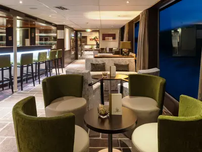 Eine moderne Bar-Lounge auf der AMADEUS Diamond. Im Vordergrund sind grüne Sessel und kleine Tische zu sehen, dahinter eine lange Bar mit Barhockern. Grosse Fenster geben den Blick auf dunkles Wasser frei.