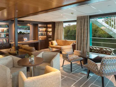 Elegante Panorama-Lounge auf der AMADEUS Diamond. Zahlreiche Bücher, ein Fernseher und bequeme Sitzgelegenheiten mit Blick auf bewaldete Flussufer.