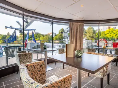 Innenansicht einer modernen Panorama-Lounge an Bord der AMADEUS Diamond. Mehrere Tische und gemusterte Sessel sind zu sehen. Große Fensterfronten bieten einen weiten Blick auf das grüne Ufer, städtische Gebäude und das Flusswasser. 