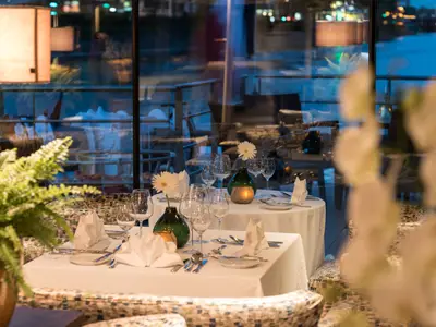 Ein elegantes Restaurant auf der AMADEUS Diamond am Abend. Die Tische sind weiss gedeckt, mit Gläsern, Besteck, gefalteten Servietten und kleinen weissen Blumen in dunklen Vasen. Kerzen in goldenen Haltern sorgen für warmes Licht. Durch grosse Fenster ist der dunkle Aussenbereich und die Reflexionen des Innenraums sichtbar.