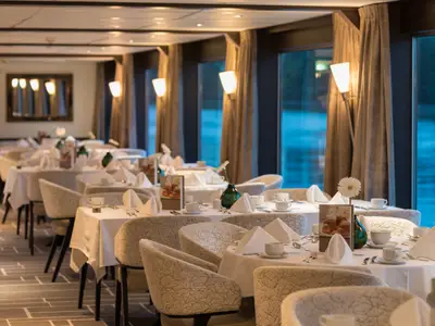 Ein elegantes Restaurant auf der AMADEUS Diamond mit vielen weiss gedeckten Tischen, bereit für Gäste. Grosse Fenster bieten einen Blick auf blaues Wasser.
