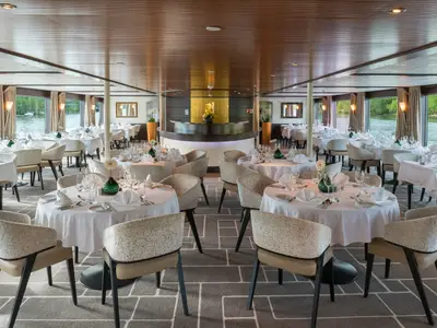 Helles, elegantes Restaurant auf der AMADEUS Diamond mit vielen runden Tischen, weißen Tischdecken, Besteck und Gläsern. Große Fenster bieten Blick auf grünes Ufer und Wasser. 
