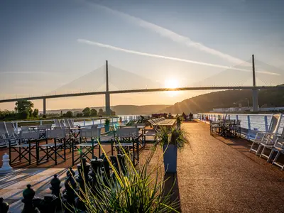 Sonnendeck der AMADEUS Diamond im Sonnenuntergang mit leeren Stühlen und Tischen. Eine große Brücke überspannt den Fluss im Hintergrund. Schachfiguren im Vordergrund.