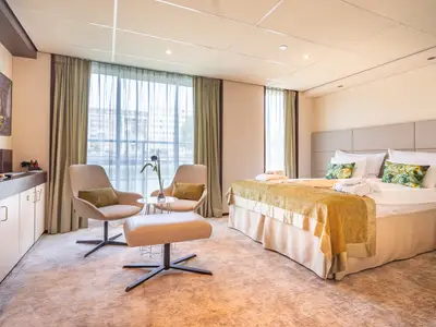 Helle, moderne Suite auf der AMADEUS Diamond mit einem grossen Doppelbett mit gruenen Kissen, einer Sitzecke mit zwei Sesseln und einem Hocker, und einem Fenster mit Blick auf eine Stadt und Wasser.