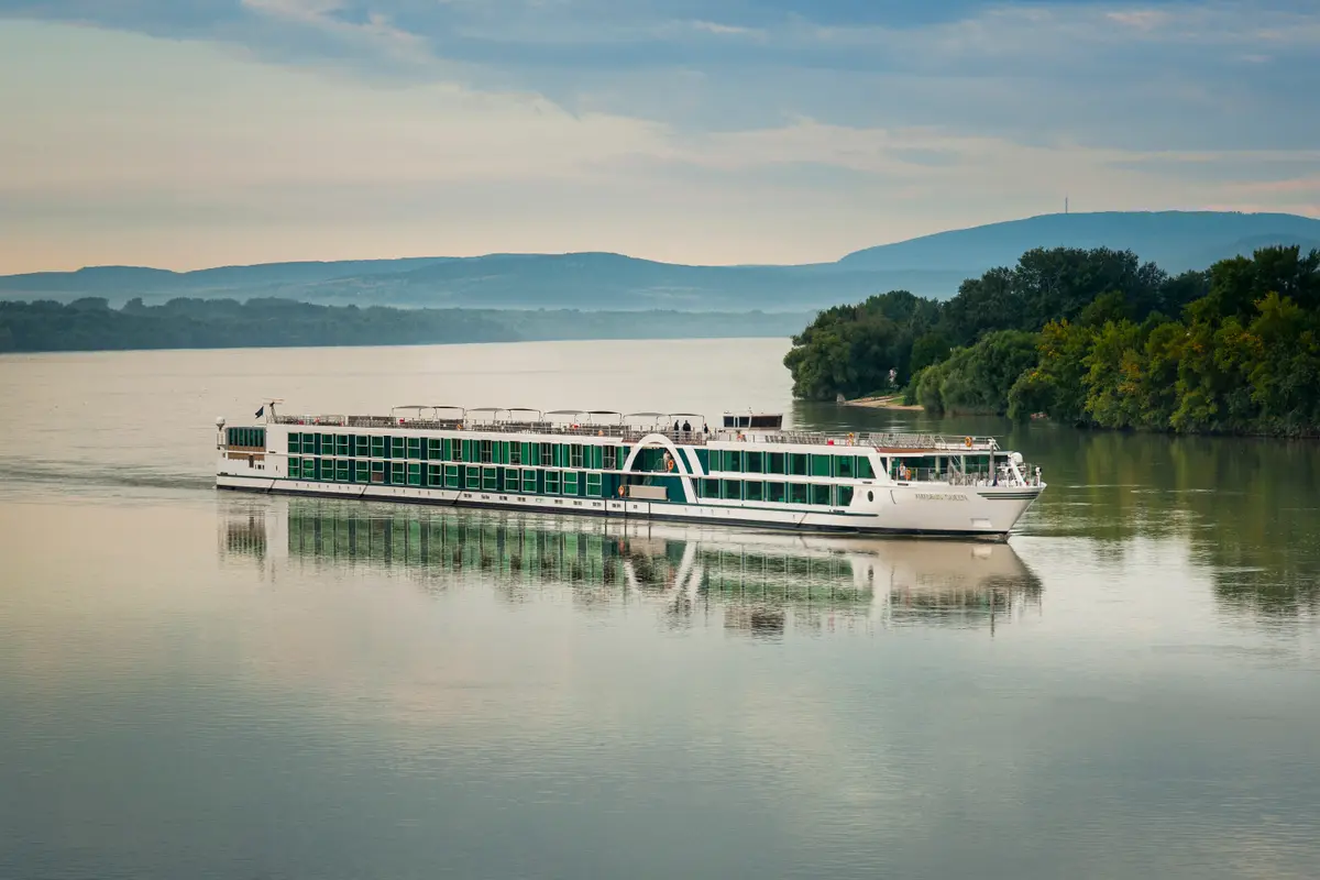 Die AMADEUS Imperial mit grünen Fenstern und Dachterrassen fährt auf einem ruhigen Fluss. Am Ufer sind dichte grüne Bäume und im Hintergrund sind sanfte Hügel und Berge unter einem bewölkten Himmel zu sehen.