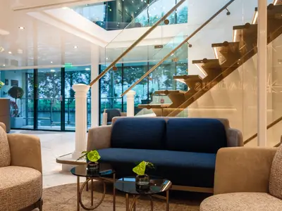 Eine moderne Lounge an Bord der AMADEUS Imperial mit bodentiefen Fenstern, die einen Blick auf Bäume und Wasser freigeben. Eine elegante Treppe mit Glasgeländer führt nach oben. Bequeme Sofas und Sessel in Blautönen und Beige laden zum Verweilen ein. Weiße Säulen und Pflanzen ergänzen das stilvolle Ambiente.