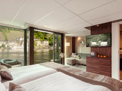 Eine moderne Balkonkabine auf der AMADEUS Imperial mit zwei Einzelbetten und einem Sofa, das sich zu einem Balkon mit Blick auf einen Fluss und eine Stadt oeffnet. An der Wand haengt ein Fernseher.