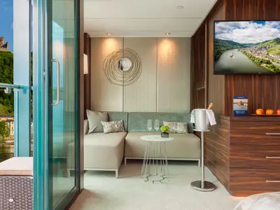 Elegante Suite auf der AMADEUS Imperial. Helles Sofa, Beistelltisch, Sektkühler. Holzschrank mit TV und Obst. Panoramablick durch Glastür auf Fluss, Berge, Burg.