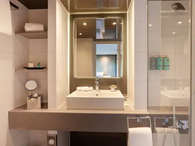 Modernes, hell gefliestes Badezimmer auf der AMADEUS Imperial mit rechteckigem Waschbecken, Armatur, einem beleuchteten Spiegel und offenen Regalen. Drei Spender sind in der Dusche sichtbar.