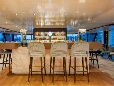 Eine moderne Bar-Lounge auf der AMADEUS Imperial mit Marmortheke, eleganten Samt-Barhockern und einer gut bestückten Bar. Große Fenster mit Blick in die Abenddämmerung.