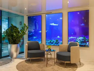 Eine moderne Lounge auf der AMADEUS Imperial mit zwei bequemen Sesseln, einem Beistelltisch und drei grossen digitalen Bildschirmen, die lebendige Unterwasserwelten mit Fischen und Korallen anzeigen. L