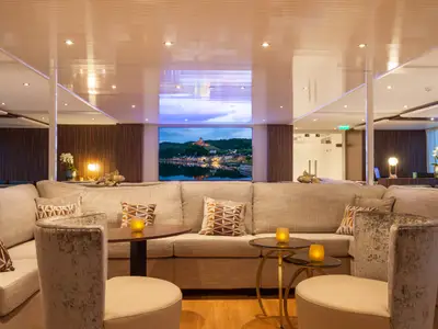 Geräumige, elegant beleuchtete Lounge der AMADEUS Imperial mit mehreren hellen Sofas und Tischen. Ein großer Bildschirm zeigt eine Stadtansicht am Fluss mit Burg, reflektiert im Wasser. Die Decke ist teilweise verspiegelt, mit vielen Einbaustrahlern.