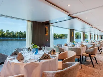 Ein elegantes Restaurant auf der AMADEUS Imperial mit vielen runden Tischen, die mit weissen Tischdecken, Gedecken und gefalteten Servietten gedeckt sind. Grosse Fenster bieten einen weiten Blick auf einen Fluss und gruene Baeume am Ufer unter blauem Himmel.