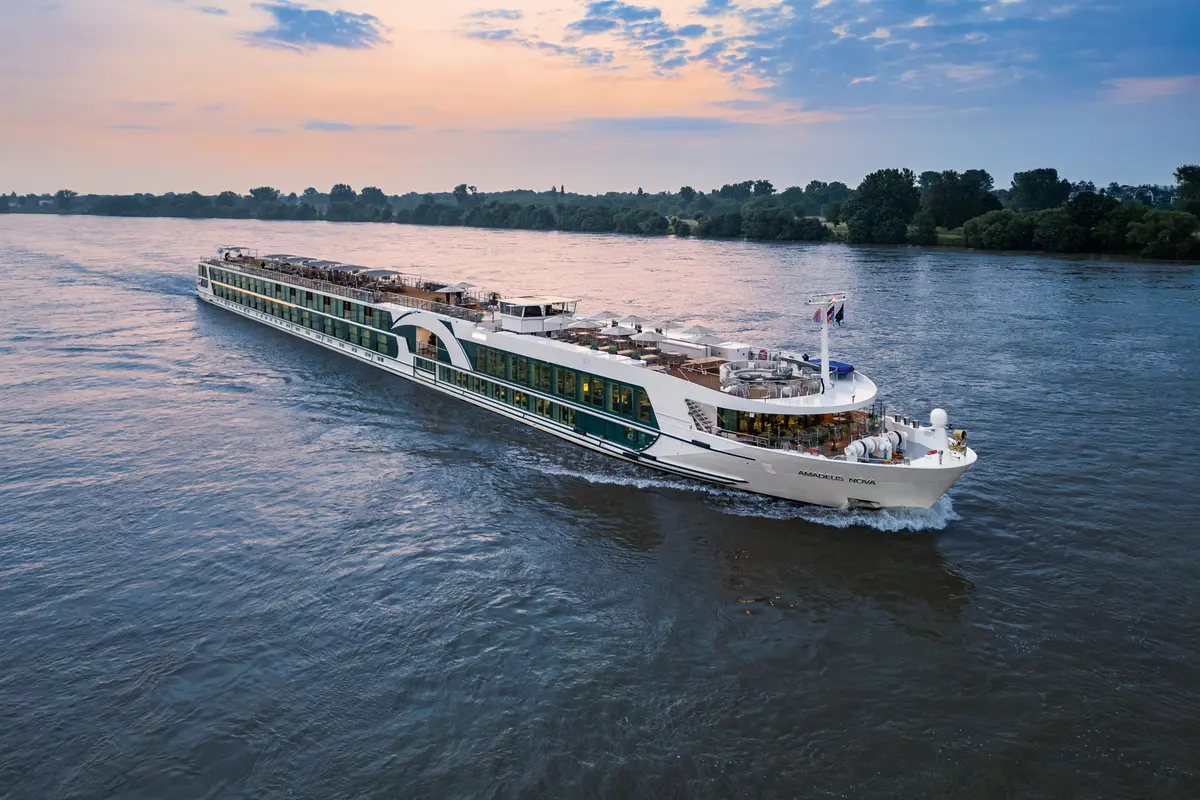 Die AMADEUS Nova, ein weiß-grünes Flussschiff, fährt auf einem breiten Fluss. Im Hintergrund säumen üppige Bäume das Ufer unter einem farbigen Abendhimmel.