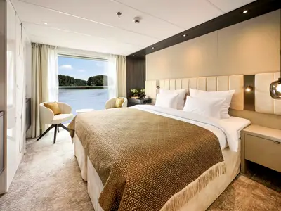 Moderne Flusskabine auf der AMADEUS Nova auf dem Mozart and Strauss Deck mit Doppelbett, Sessel und Panoramafenster. Blick auf Fluss und bewaldete Ufer. Elegante Einrichtung in Beige.